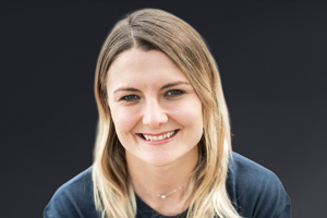 Porträt von Iris Schmiedbauer mit lächelndem Gesicht und blonden Haaren vor dunklem Hintergrund.